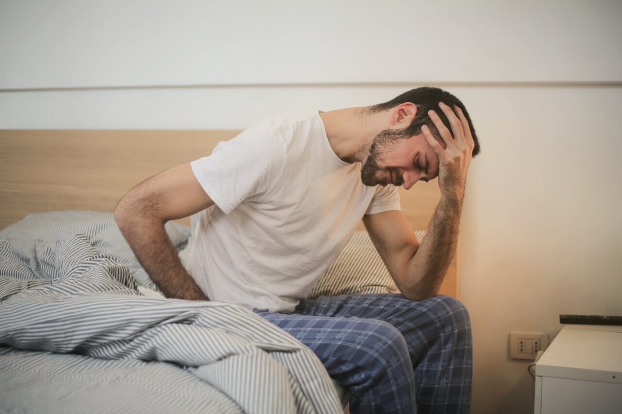 A tired man sitting on the edge of his bed with a headache.